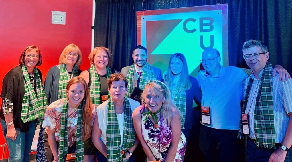 Group of ten people huddled with each other smiling at the camera, all wearing green plaid scarfs.